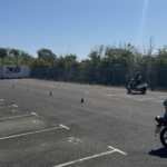 Riders on motorcycles in a large paved lot marked with white lines and cone markers, with a '2Moto' container in the background.