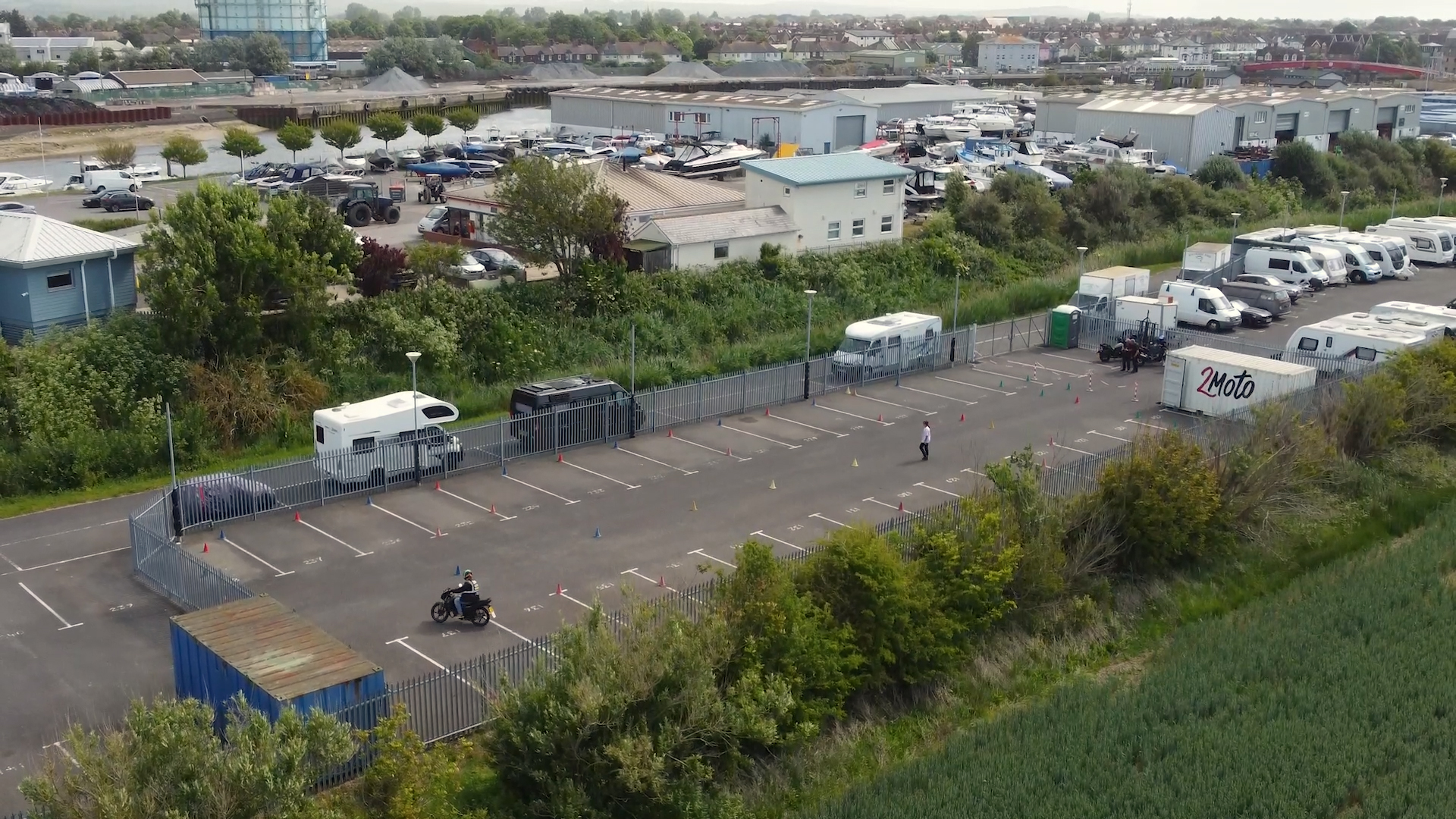 Aerial view of a fenced parking lot with caravans, cars, and a motorbike; cones mark parking spaces near greenery and buildings in the background.