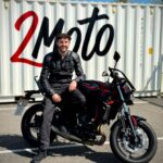 Smiling man in black leather riding gear sits on a black and red Kawasaki motorcycle in a parking lot with a large white container behind him.