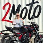 Woman in sunglasses holding two papers beside a black and red Kawasaki motorcycle in front of a wall with large 'Lotto' graffiti.